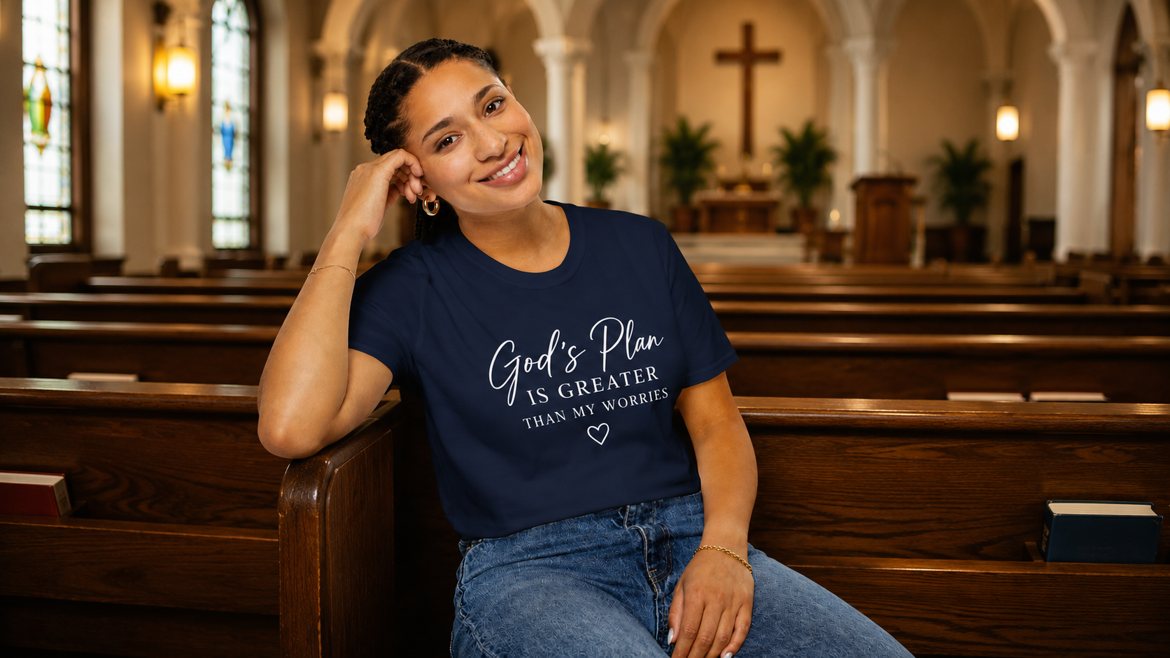 A woman sitting in a wooden church pew wearing a navy blue graphic t-shirt that reads 'God's Plan IS GREATER THAN MY WORRIES' in white script, with a heart logo underneath.